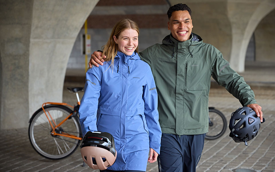 Lachend duo in blauwe en groene jas met fietshelmen in een overdekte doorgang.